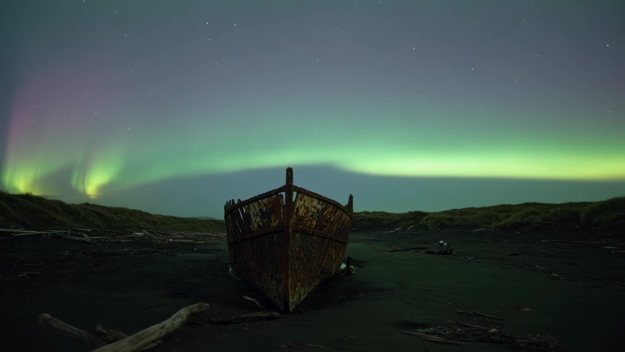 Et lysende nordlys-bånd bevæger sig over en rusten strandet båd på sort sand, med en reflekterende vandpyt.