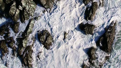 slow motion ocean waves and white foam crashing over cornish rocks with an aerial top down shot