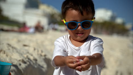 Zeitlupe-Eines-Süßen-Jungen-Mexikanischen-Lateinamerikanischen-Jungen-Mit-Sonnenbrille-Und-Weißem-T-Shirt,-Der-Der-Kamera-Einige-Muscheln-Zeigt-Und-Sie-Dann-In-Einen-Eimer-Legt