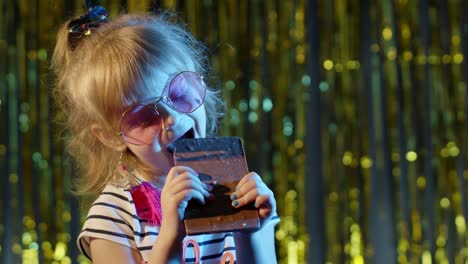 stylish trendy young child girl at disco party cyberpunk club eating big chocolate bar sweet candy