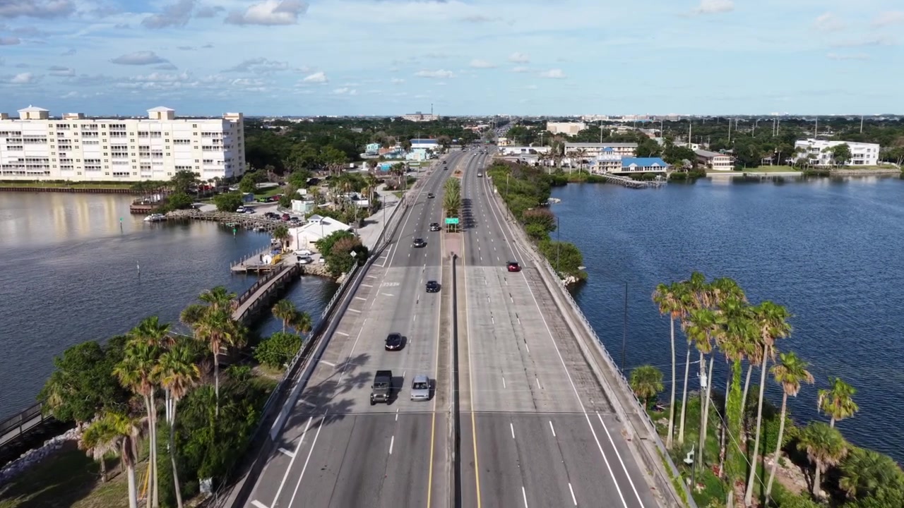 Ponte a carreggiate separate che trasporta auto sotto uno spartitraffico fiancheggiato da palme verso una città distante, con un tranquillo corso d'acqua blu che fiancheggia la carreggiata su entrambi i lati vicino a Merritt Island, costa della Florida. Ampia ripresa aerea con drone