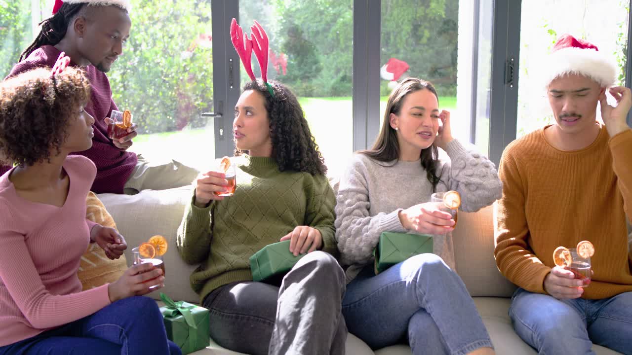 Diverse friends exchanging gifts and toasting drinks with standing friend joining in living room