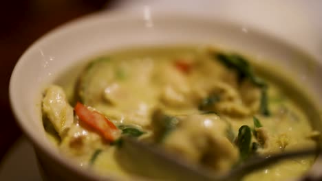 a spoon stirs vibrant thai green curry, revealing colorful vegetables and chicken in a creamy sauce under warm lighting