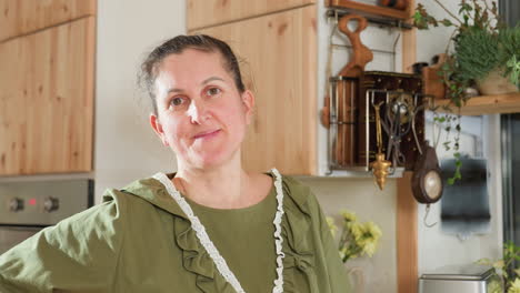 portrait view of middle-aged lady turning to camera with soft smile and blinking eyes while standing confidently in warm, rustic kitchen setting filled with wooden cabinets, utensils, and plants