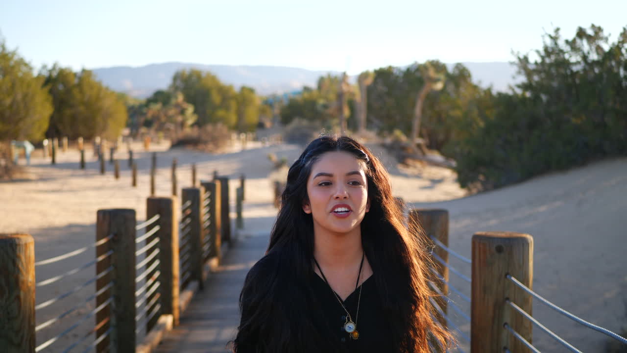 una chica guapa con el pelo largo y negro sonriendo alegremente mientras camina por un puente de madera del desierto en cámara lenta al atardecer