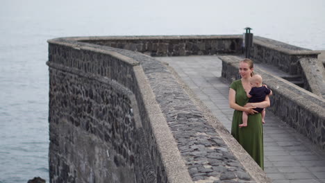 enjoying the atmosphere of an ancient european square by the ocean, a young mother walks with her baby son, gazing at the waves and sharing smiles