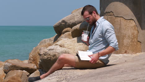 Man by the ocean study reading a book and enjoying cold drink