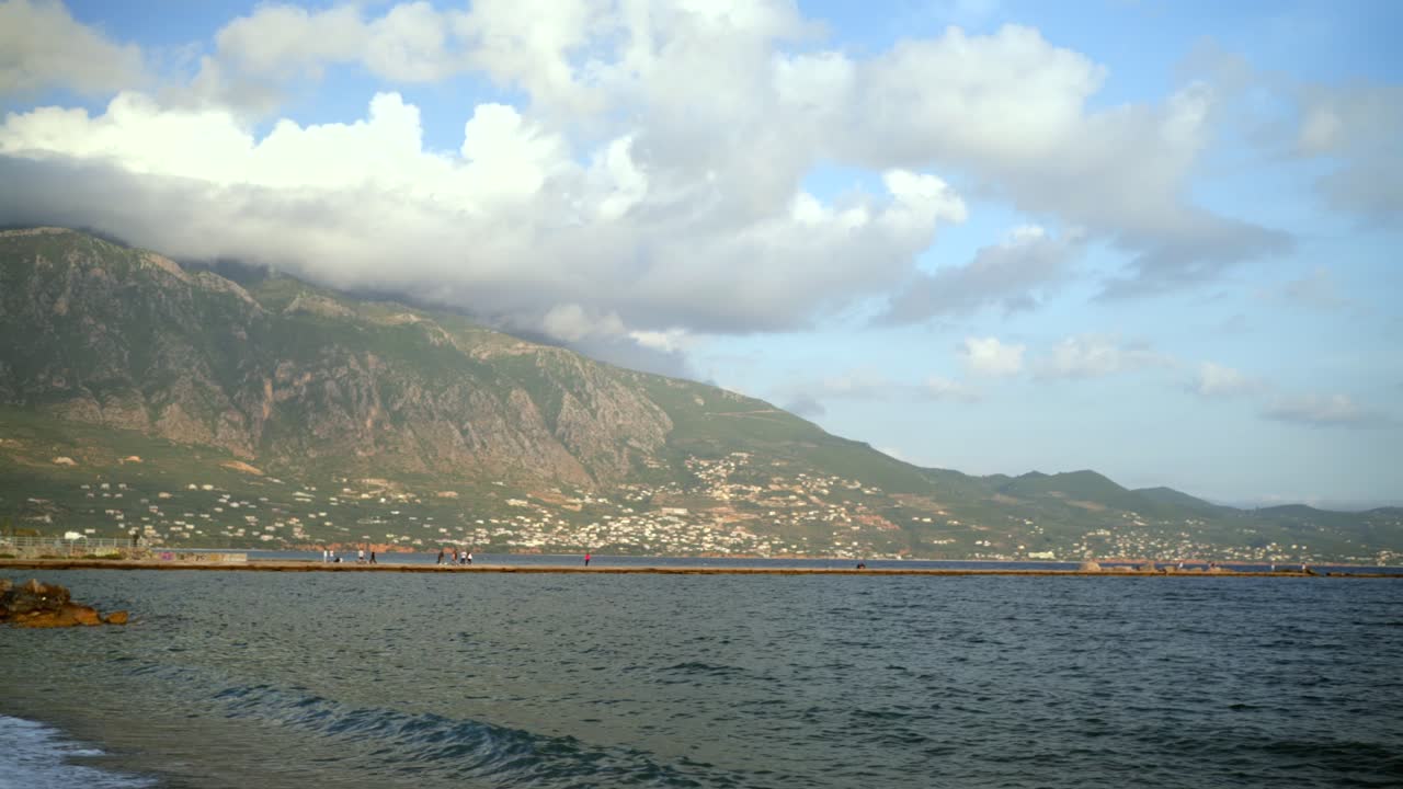 Wide panoramic view of sea of Kalamata, people walk on Dark Pier, Taygetos mountain on background covered with clouds 4K