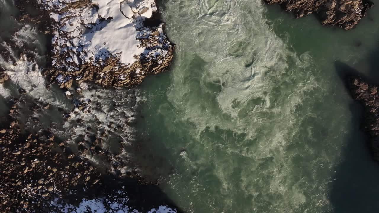 Top-down aerial view of Urriðafoss waterfall in Iceland, showcasing turbulent glacial waters flowing through rugged, snow-covered volcanic rocks. Capturing the raw power of nature.