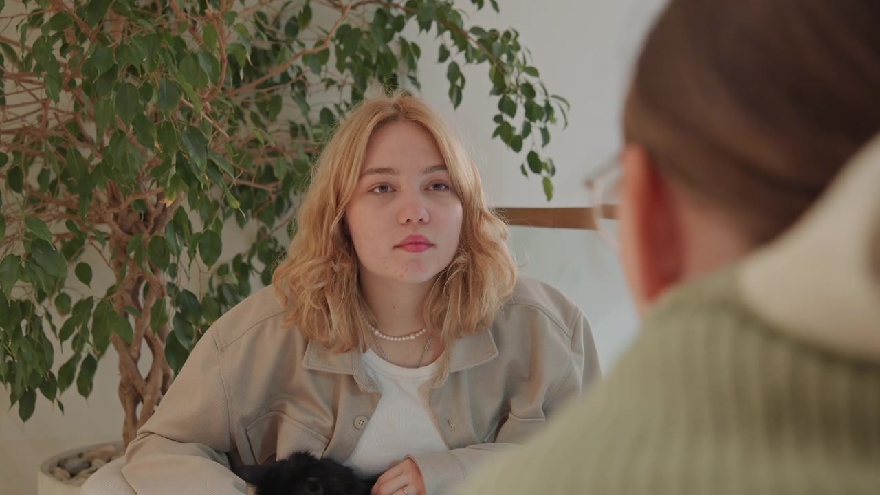 Close up of woman with blonde hair holding and petting black cat while sitting in indoor setting. scene features soft lighting natural textures like plant background and calm interaction