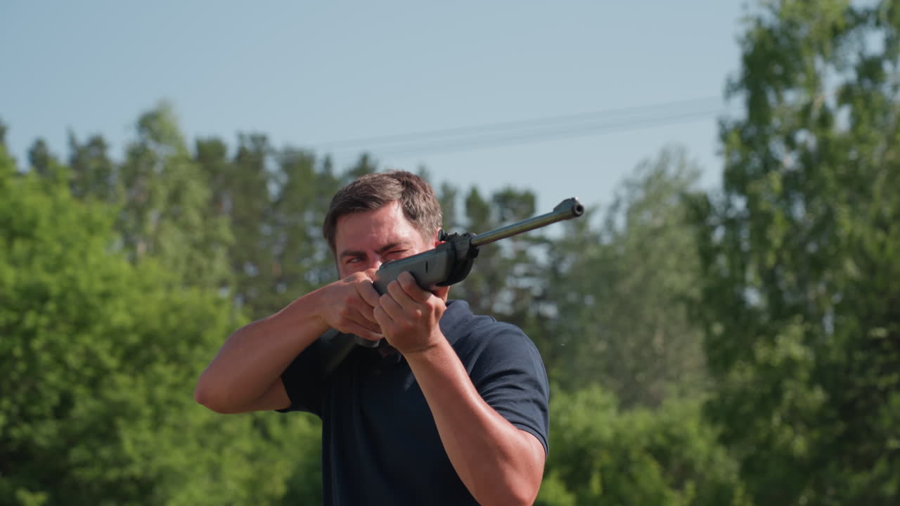 man aiming rifle into distance then lowering weapon while wiping ear from sweat, tense focus on rural property with green trees background, protective pest control action