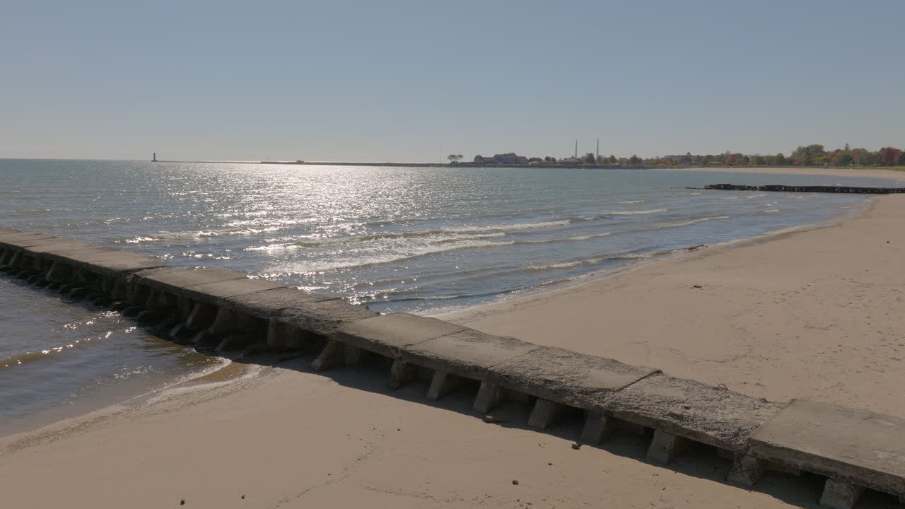 Cinematic closeup of an old concrete pier on the beach with a pan revealing it stretching into Lake Michigan, sunlight shimmering across the water on a pretty day