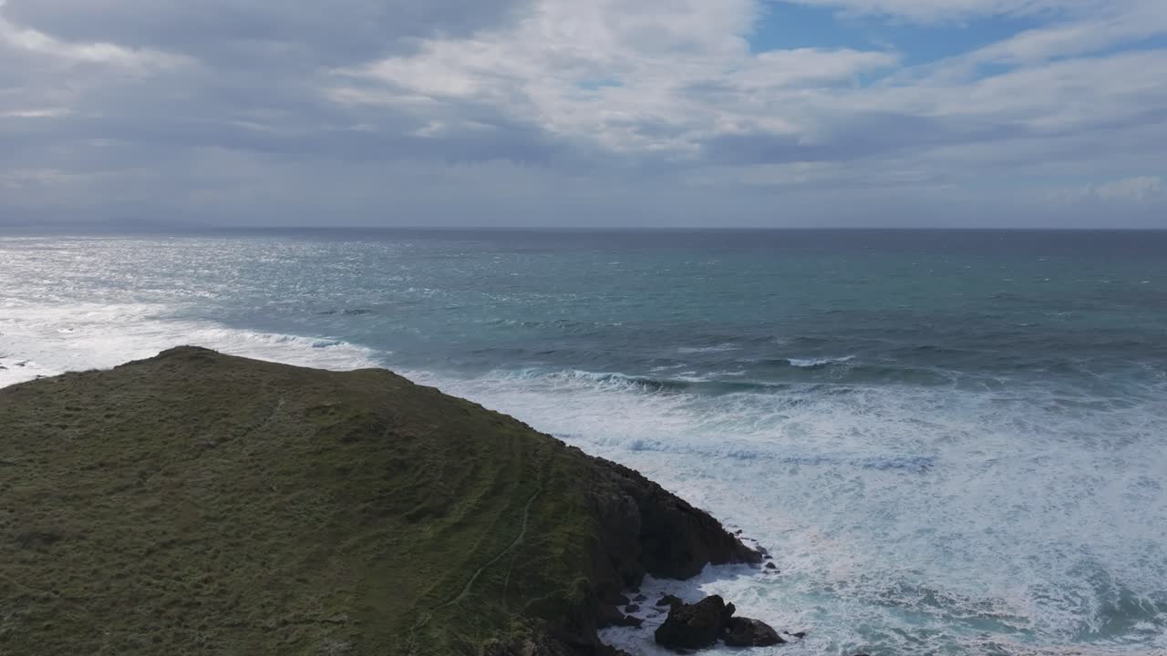 Lateral aerial shot along the Cantabrian coast showing rough waves crashing against a small green peninsula, creating white foam. Storm clouds gather above, highlighting the dramatic coastal landscape