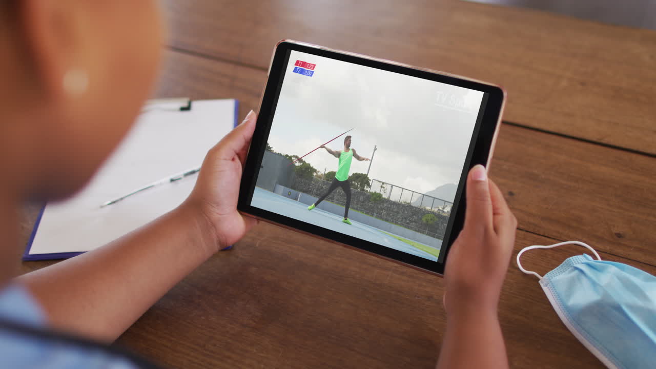 태블릿에서 육상 자벨린 경기를 시청하는 얼굴 마스크와 함께 테이블에 앉아있는 여성의 복합