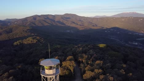 Three-runners-approach-tall-watch-tower-in-Tordera-trailrace-at-sunset-with-brown-mountain-range-in-background,-above-aerial-approach