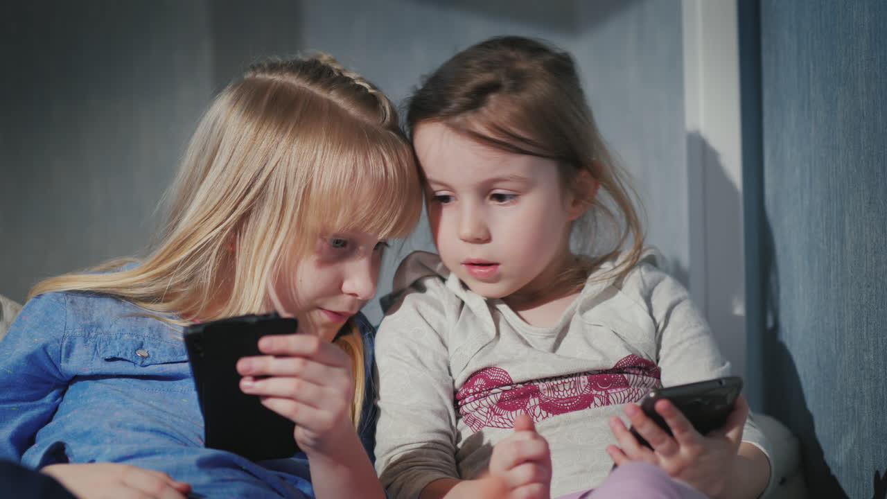 스마트폰으로 노는 두 소녀는 침실 4k 비디오에서 침대에 나란히 앉아 있습니다.
