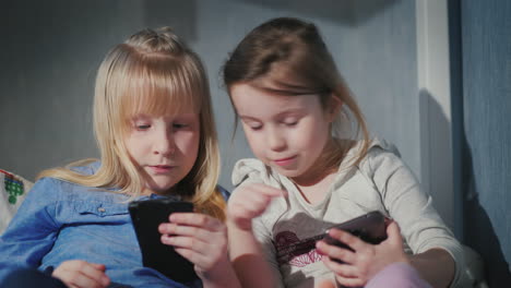 dos chicas juegan en los teléfonos inteligentes sentadas una al lado de la otra en la cama en el video de 4k de su dormitorio