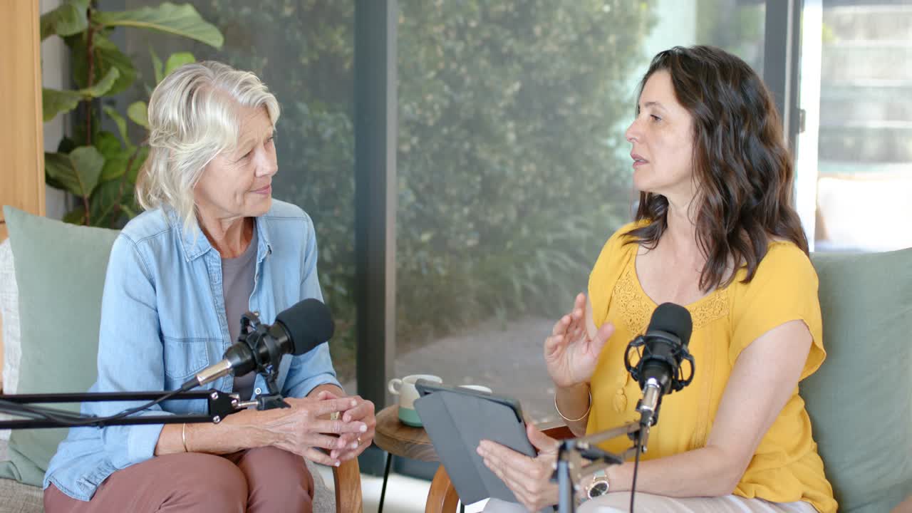 Vrouwelijke presentatrices presenteren podcast met microfoons, tablet waardoor de rechter presentatrice leest en gebaart, linker knikt.