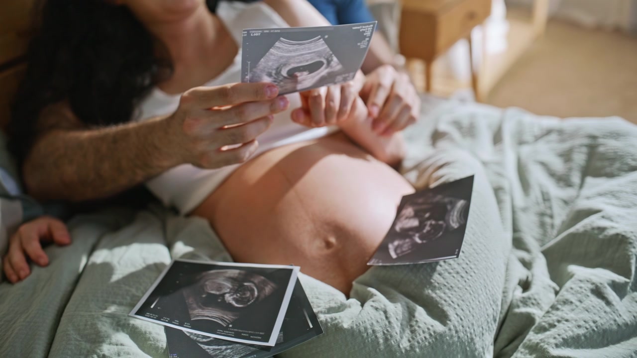 Closeup couple holding sonogram photos over pregnant belly. Ultrasound on bed.