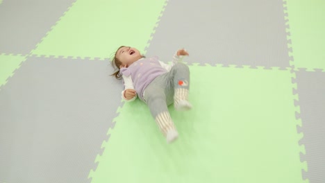 Happy-Two-year-old-Baby-Girl-Yelling-and-Shaking-Legs-From-Laughter-Lying-on-a-Checkered-Mat-Floor---slow-motion