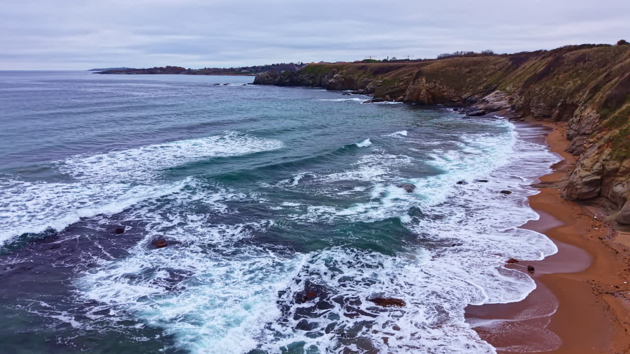 Waves crash on sandy beach at coastline with rocky cliffs during cloudy day