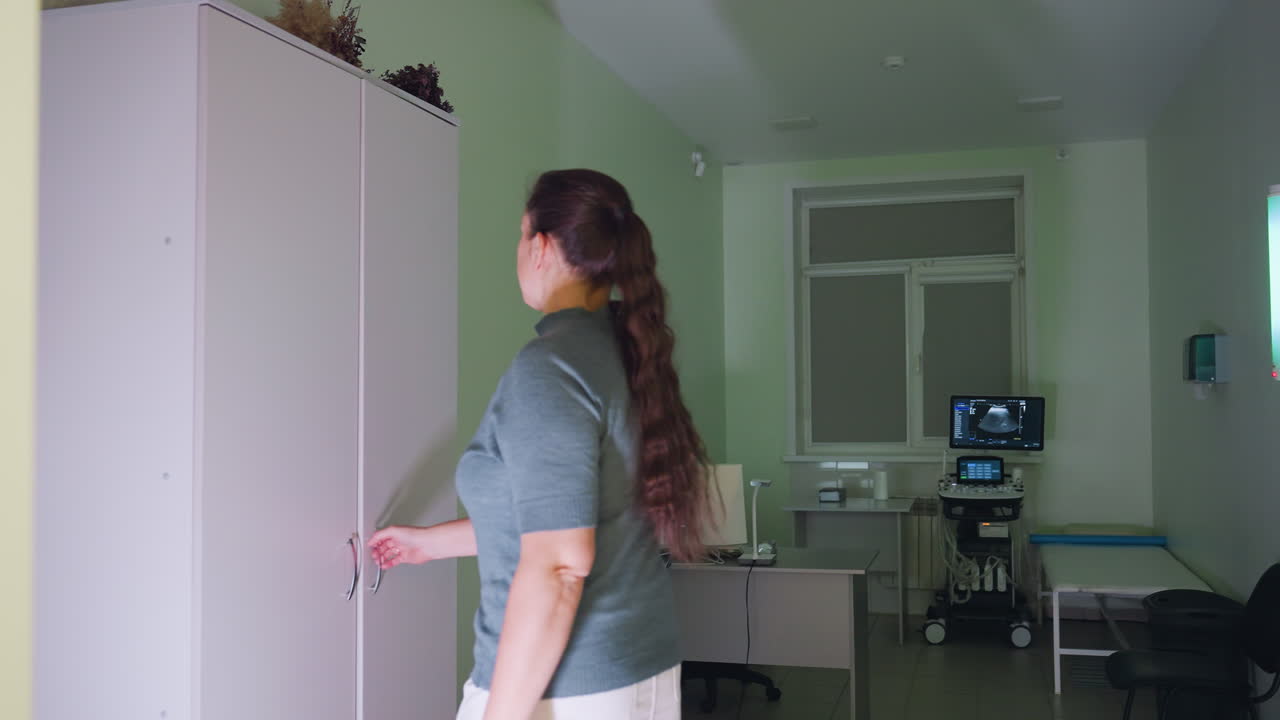 Medical personnel enters examination room walks toward locker with flower vase on top opens it to take out clothing for work preparation in clean modern clinic with equipment and green interior walls