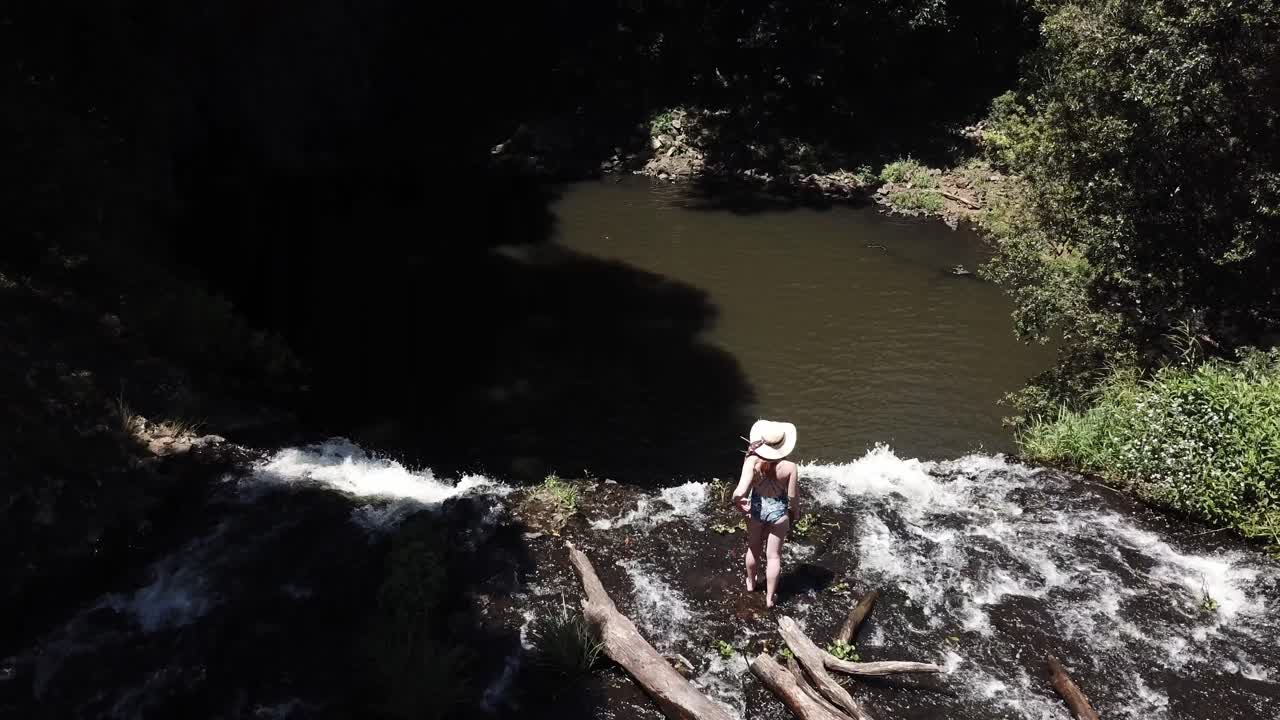 drone aéreo en la naturaleza con una mujer caminando en la cornisa de una cascada explorando con la cámara panorámica hacia abajo