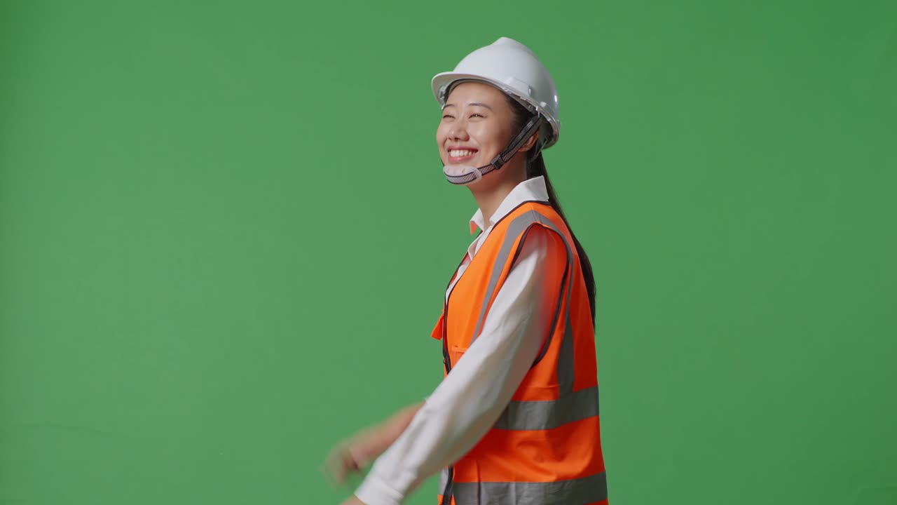 안전 헬을 입은 아시아 여성 엔지니어가 녹색 스크린 배경 스튜디오에서 는 동안 손을 흔들고 있는 측면 시각