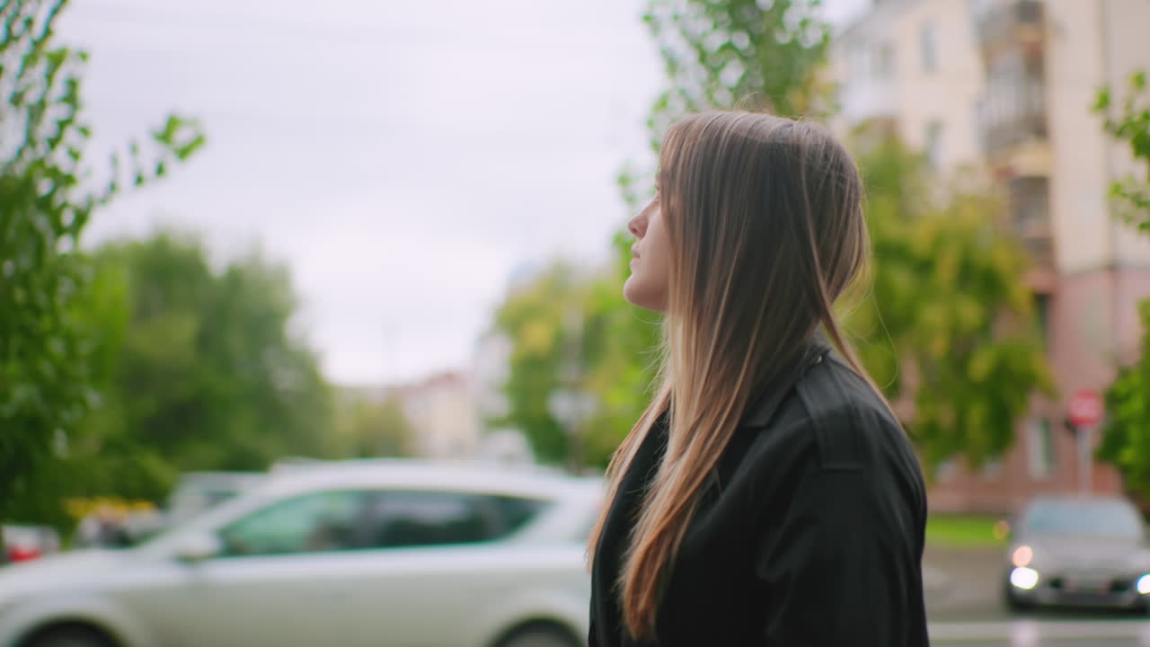 Woman wearing black coat holding phone to ear while standing outdoors in city park area with blurred cars in background, expressing concentration and seriousness during call on cloudy day