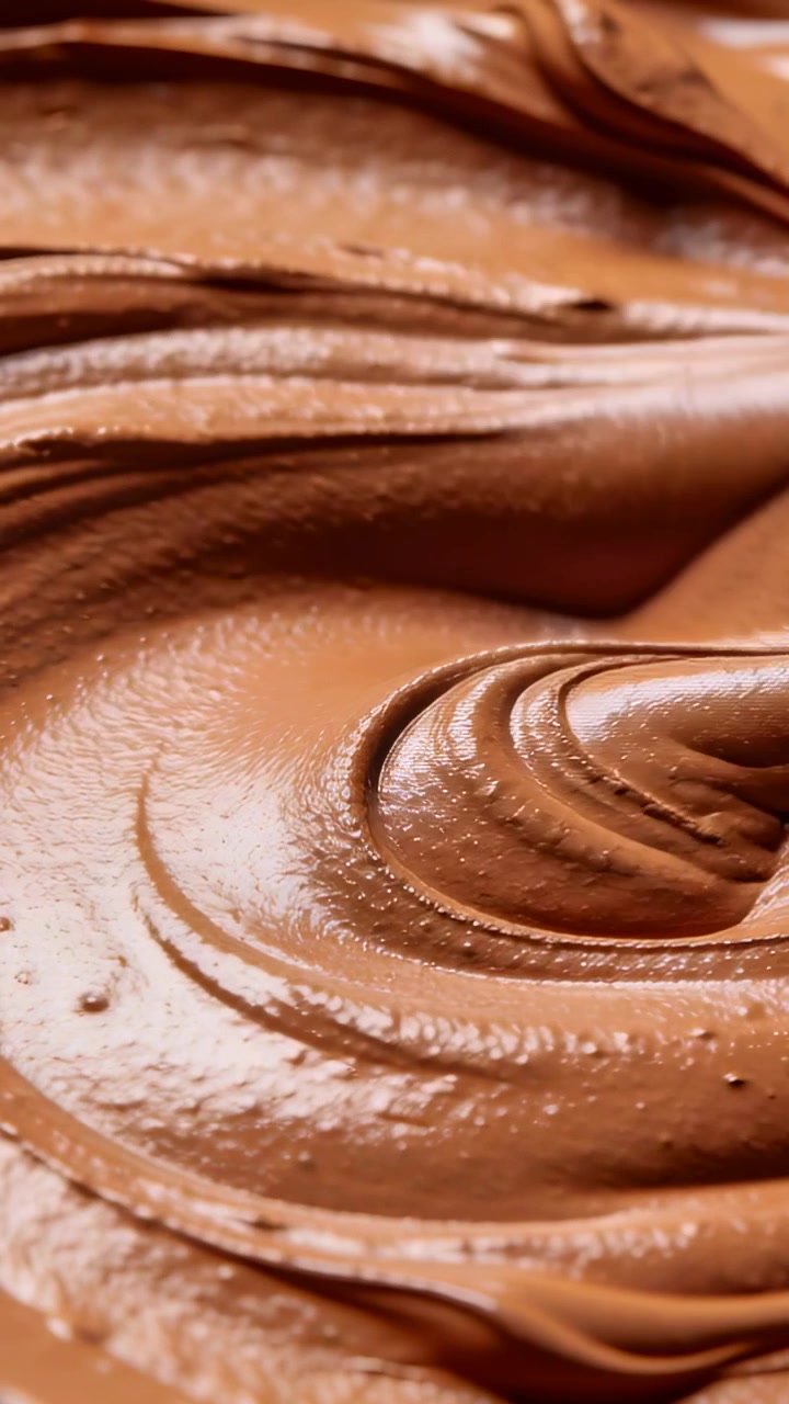 Video verticale: Spatola chiara che, provenendo da destra, amalgama cioccolato lucido in una ciotola, creando un vortice uniforme.