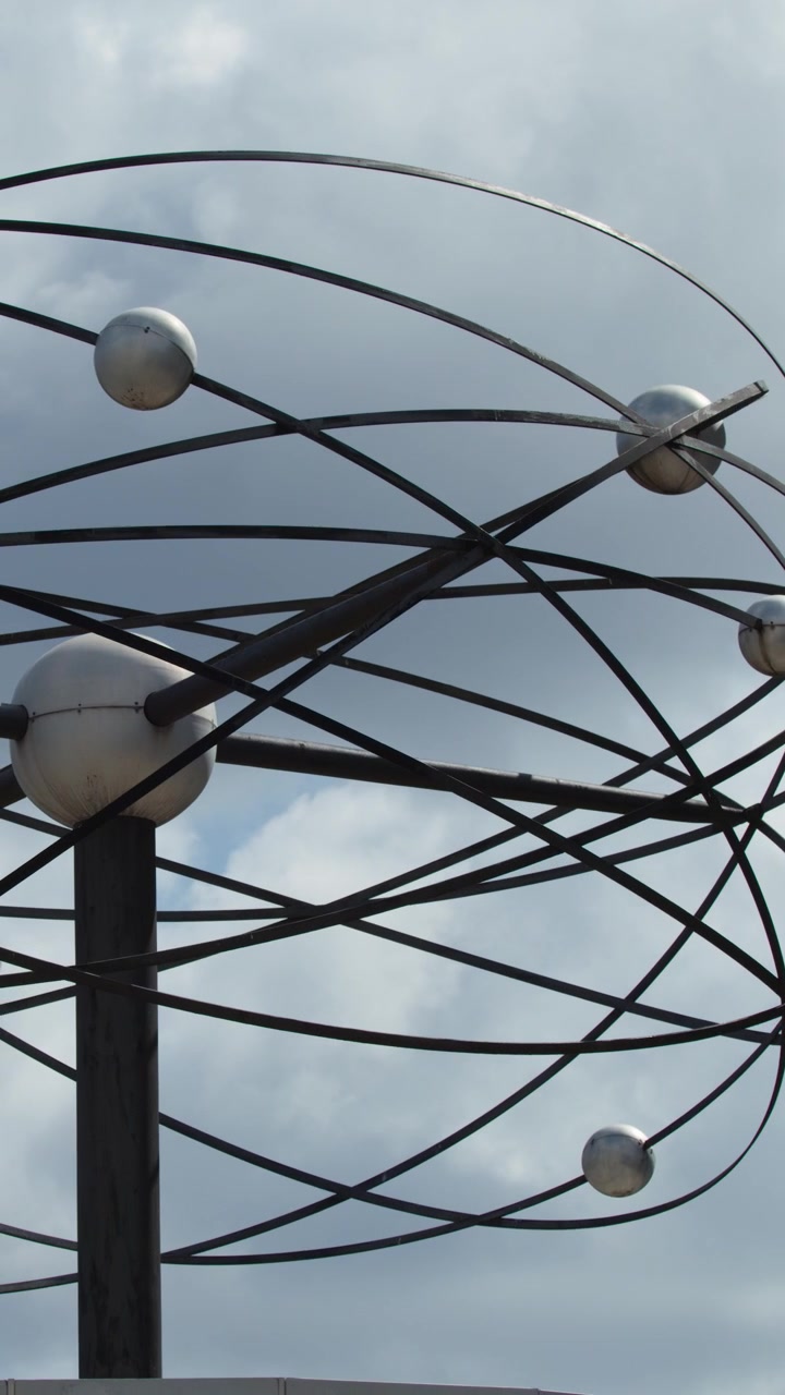 Una scultura a sfera metallica ruota contro un cielo nuvoloso, in un'ambientazione urbana, con telecamera fissa, luce naturale del giorno e focus architettonico.