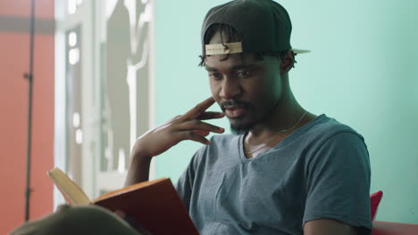 Close up of young man in casual shirt and cap reading book with focused expression, hand near face, seated indoors against turquoise wall with silhouette artwork in background