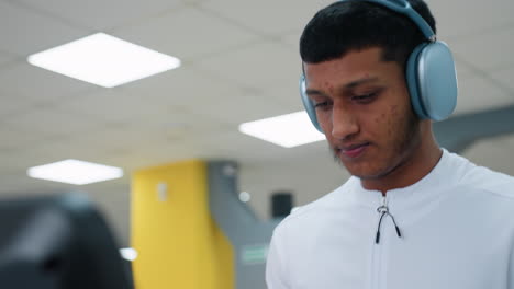focused athlete with acne wearing over ear headphones on treadmill under bright ceiling lights in gym, exercising with determination as blurred background