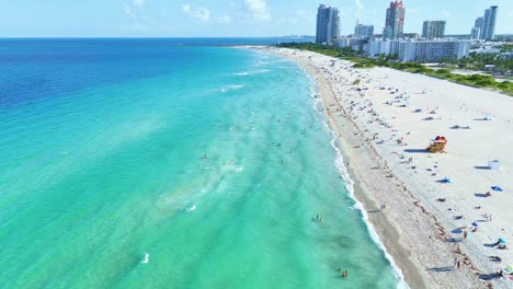 A-drone-flies-400ft-over-Miami-Beach,-showing-waves-hitting-the-shore,-people-in-the-water,-beachgoers-with-umbrellas-and-chairs,-and-a-serene-coastal-scene