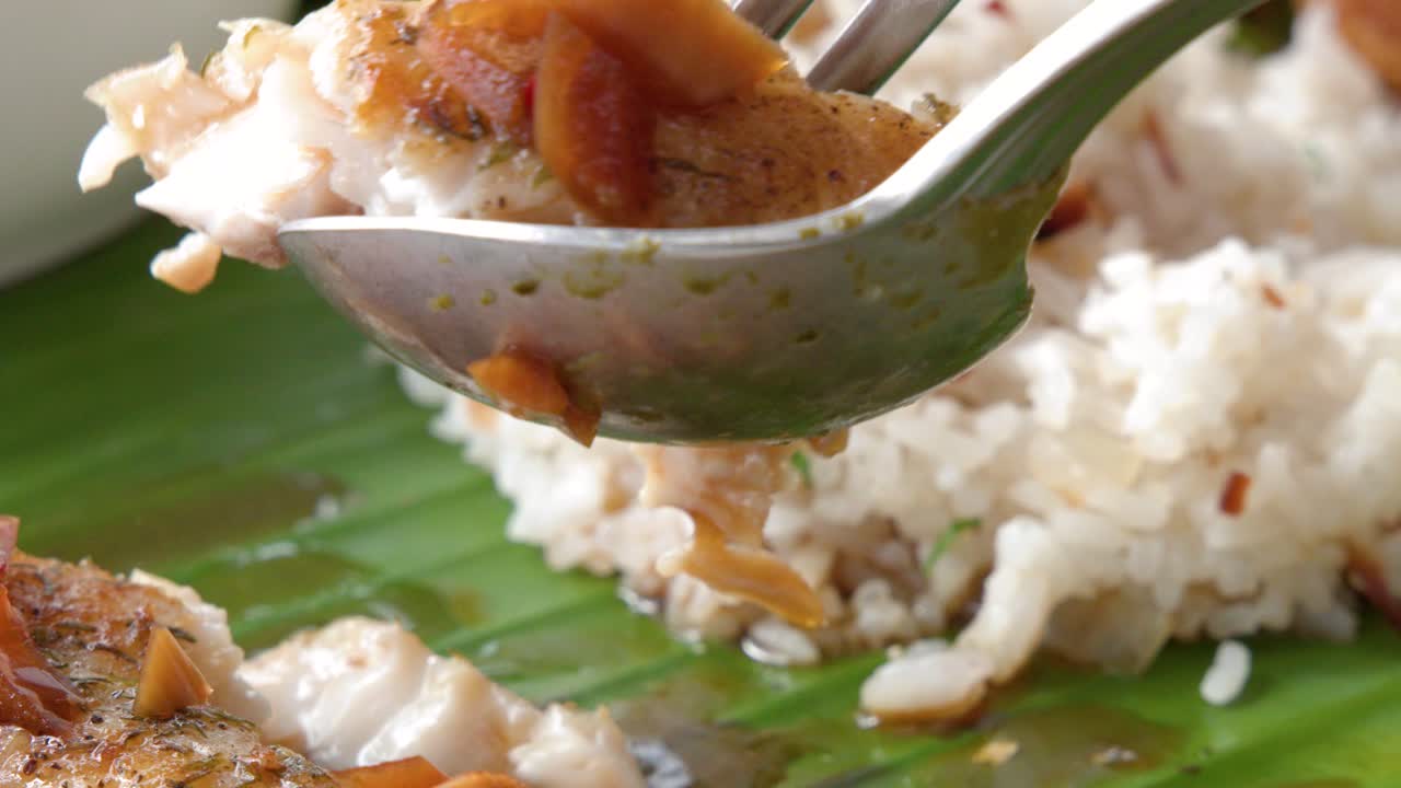 Foto close-up seseorang menggunakan sendok untuk makan ikan bakar dengan nasi putih.