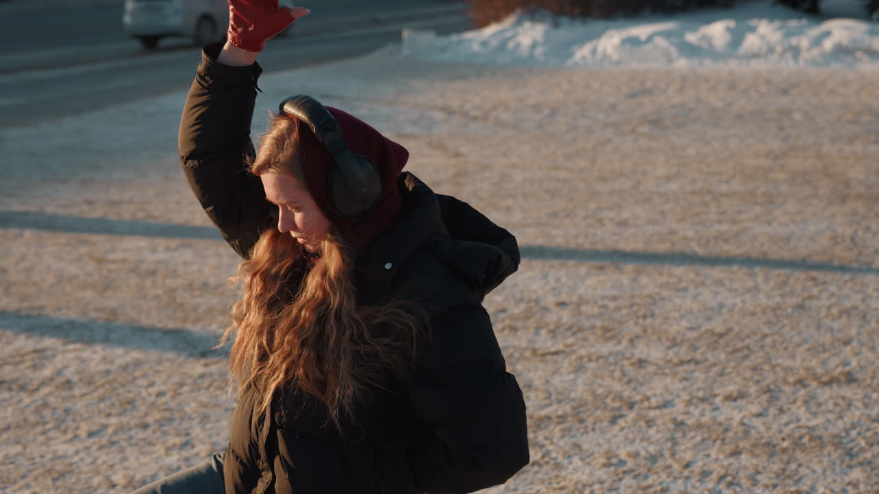 Donna in un campo innevato improvvisa passi di danza con guanti rossi e cuffie durante l'ora d'oro, capelli al vento e cappotto caldo, lunghe ombre e strada urbana sullo sfondo, umore giocoso e solitario, atmosfera cinematografica.