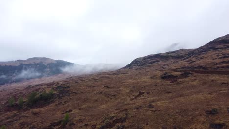 Aerial drone footage slowly flying closer and gliding over brown grassy mountain base or hillside in Scotland while white fluffy and soft clouds are low to the ground. Misty, foggy day with white sky.
