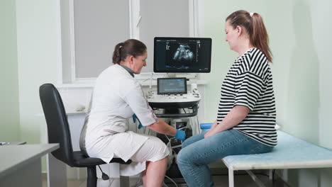 lab scientist in white coat gently touches printed ultrasound images on printer while having post-examination conversation with pregnant patient with visible monitor displaying baby scan