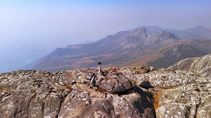 Un drone orbita attorno agli escursionisti accanto al segnavia della vetta sul Picco Sapitwa, mostrando ampie vedute montane attraverso il paesaggio aspro del Mulanje in Malawi.