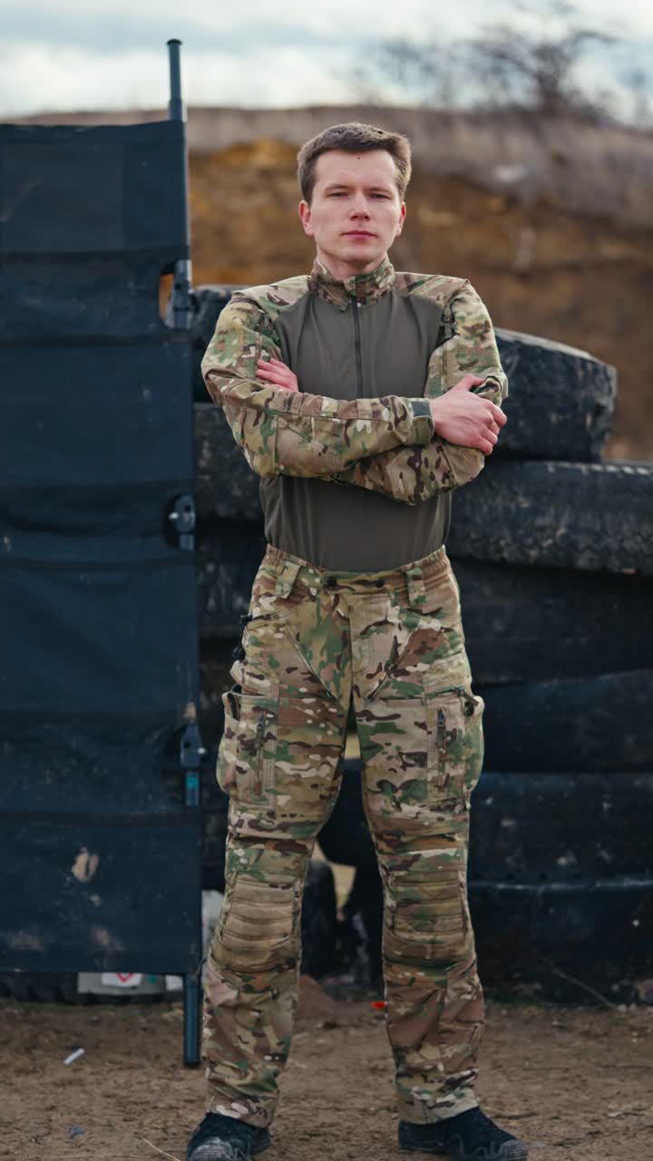 retrato vertical de video de un joven confiado en ropa de camuflaje de pie cerca de neumáticos durante ejercicios de combate en la estepa. retrato de un joven militar confiado en ropas militares modernas