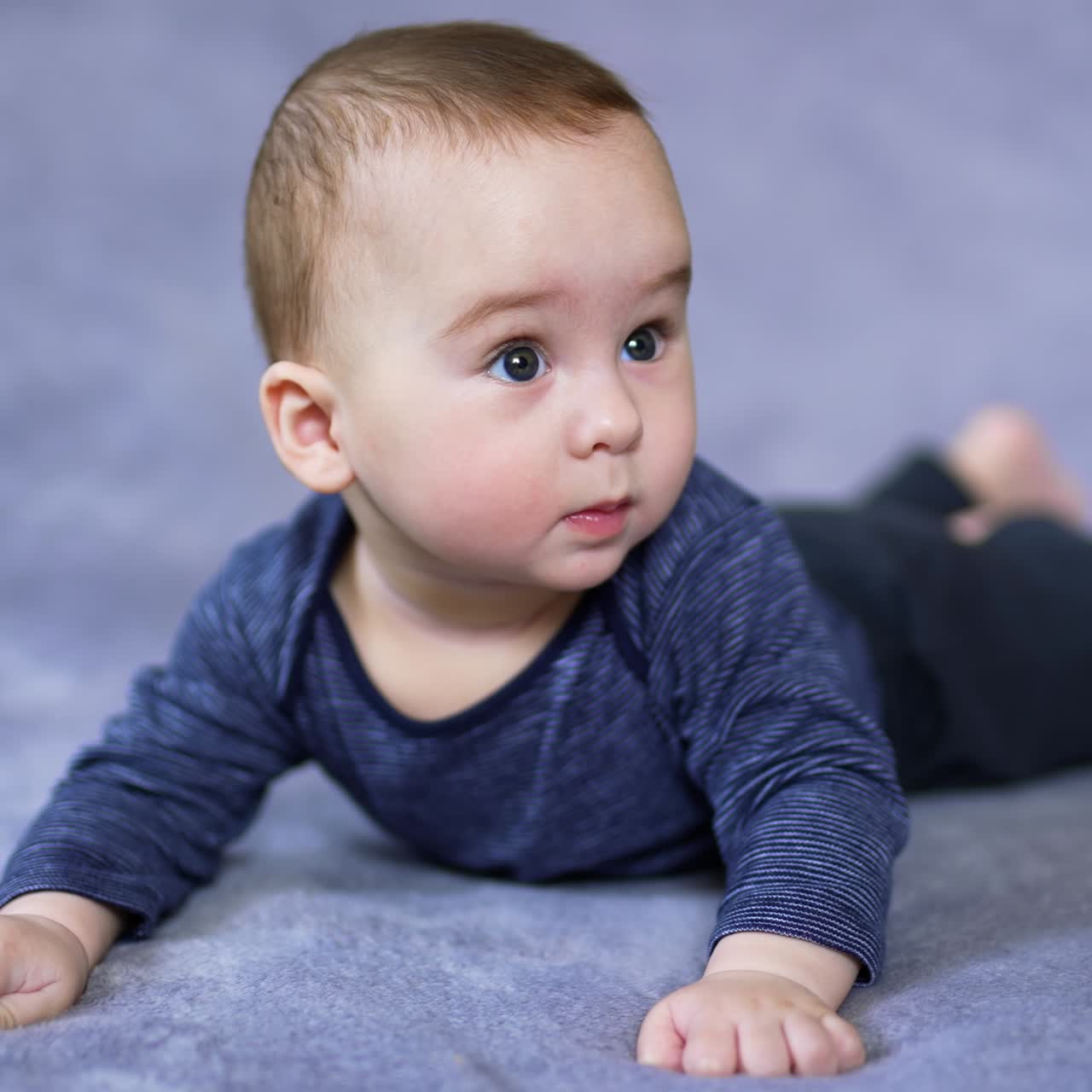 Adorable Caucasian boy in blue clothes lies on belly waving his feet. Nice curious toddler looks up with interest sweetly rising his eyebrows
