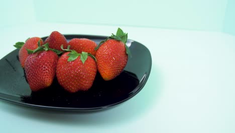 fresh big red tasty ripe strawberries rotates slowly on a black plate on light blue background, healthy food concept, medium shot, camera rotate right