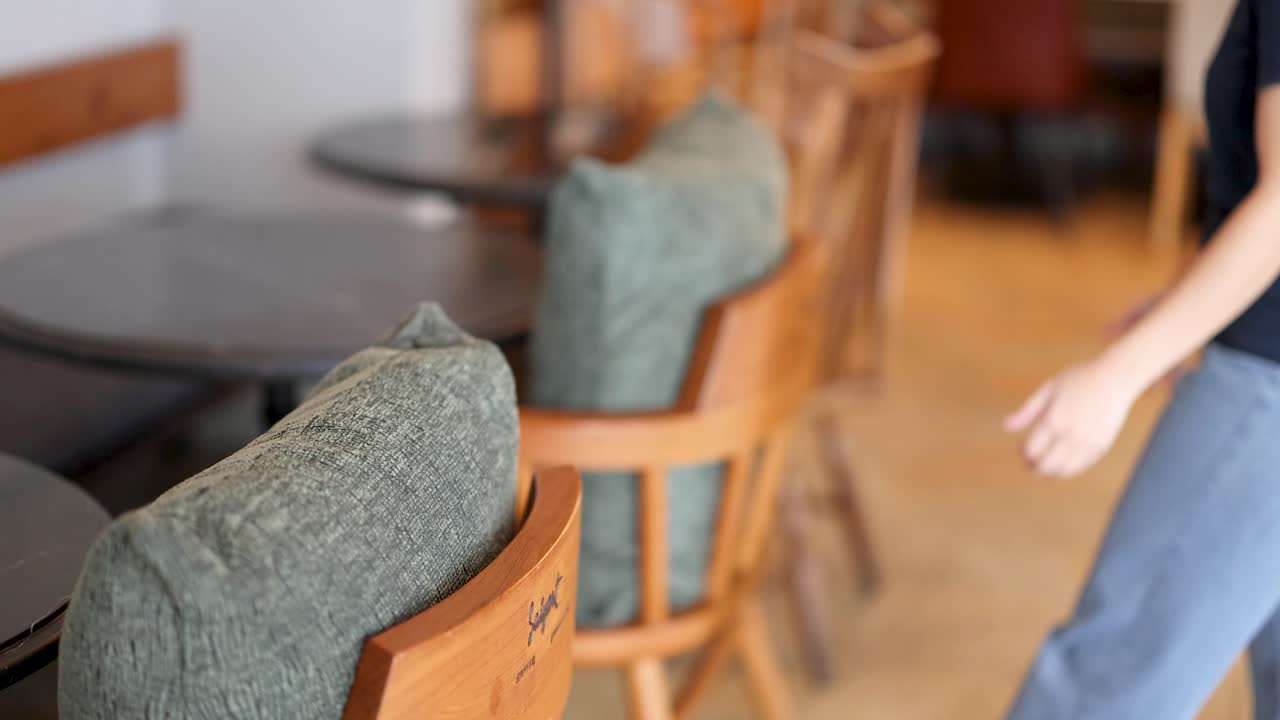 Person arranges green cushion on wooden chair in softly lit, modern cafe with wooden furniture