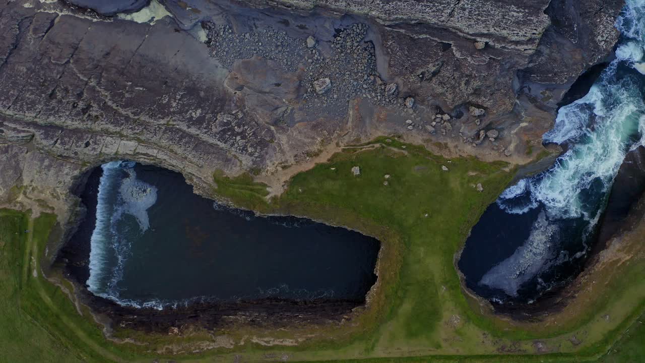 Drone top down descends on grassy plains above Bridge of Ross sea arch as waves churn in the waters below