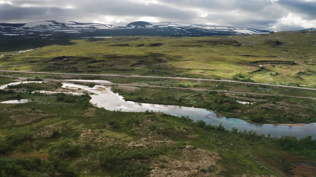 un vistazo a la naturaleza tranquila del círculo polar ártico: un río poco profundo corre tranquilamente cerca de un ferrocarril, una carretera asfaltada está al lado de una ladera verde, los picos nevados están tocando el cielo nublado