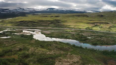 Ein-Flüchtiger-Blick-Auf-Die-Ruhige-Natur-Des-Polarkreises:-Ein-Flacher-Fluss-Fließt-Ruhig-In-Der-Nähe-Einer-Eisenbahn,-Eine-Asphaltstraße-Grenzt-An-Grüne-Hügel,-Schneebedeckte-Gipfel-Berühren-Den-Wolkigen-Himmel