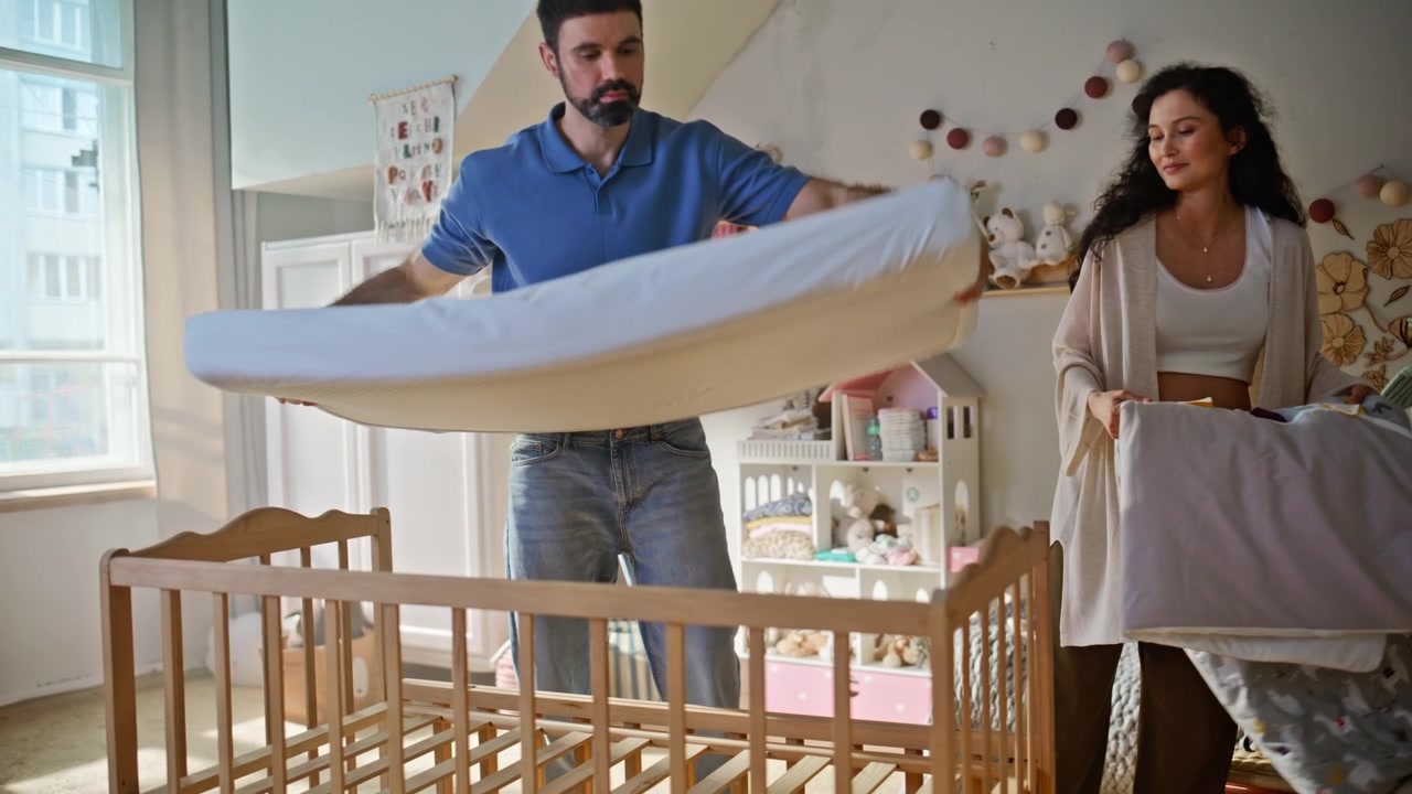 Man assembling wooden crib nursery. Pregnant wife helping place mattress home.