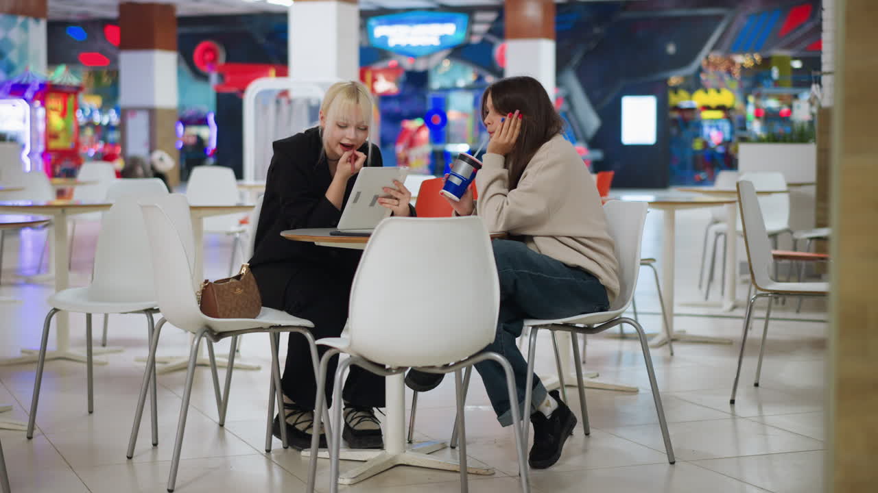 Two sisters seated in modern cafe as one applies lipstick using tablet mirror while other drinks from cup, showing contrast of focus and disinterest with colorful blurred background