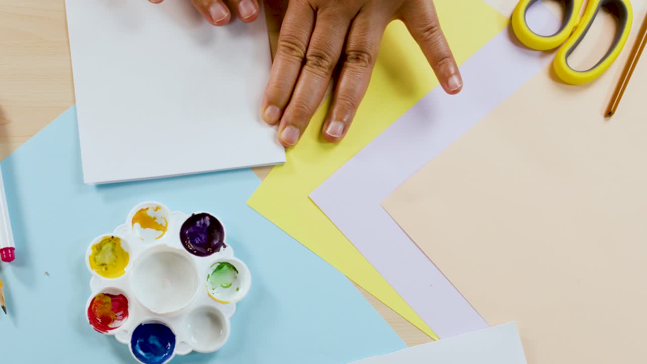 Hands fold white paper over wet paint blots on a colorful desk with art supplies