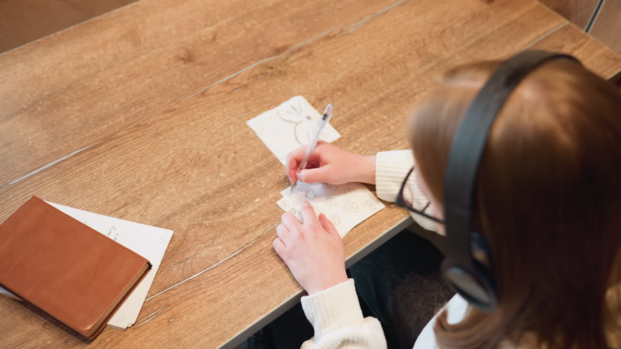 Top down view of woman with brown hair wearing headphones sketching floral patterns on tissue paper using pen while seated at wooden table with notebook and brown book beside her